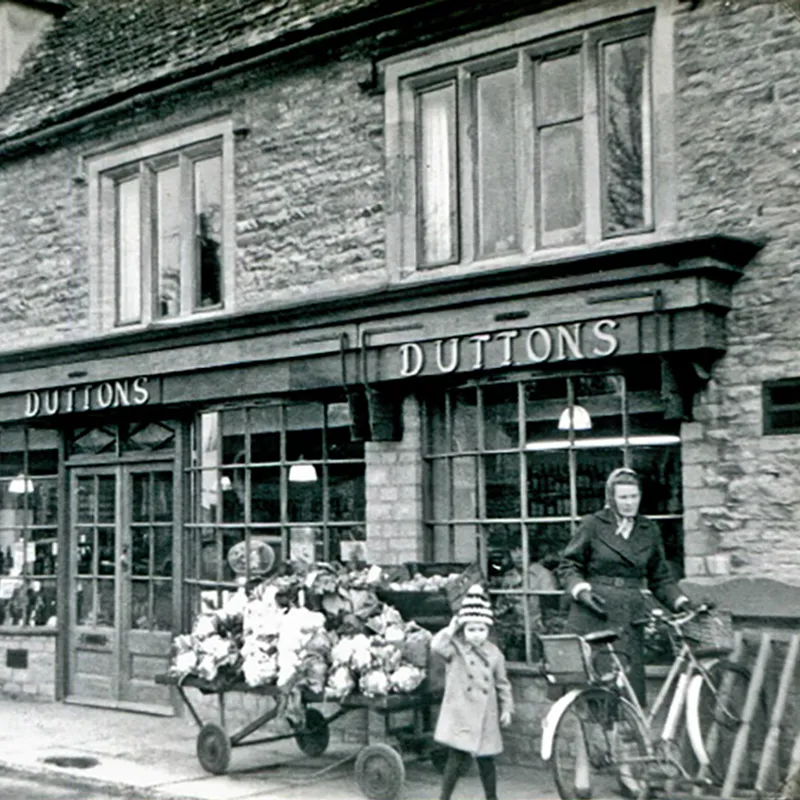 Bampton Housing & Building Black and white Duttons shop front with a lady about to get on a bike with a young child stood next to her.