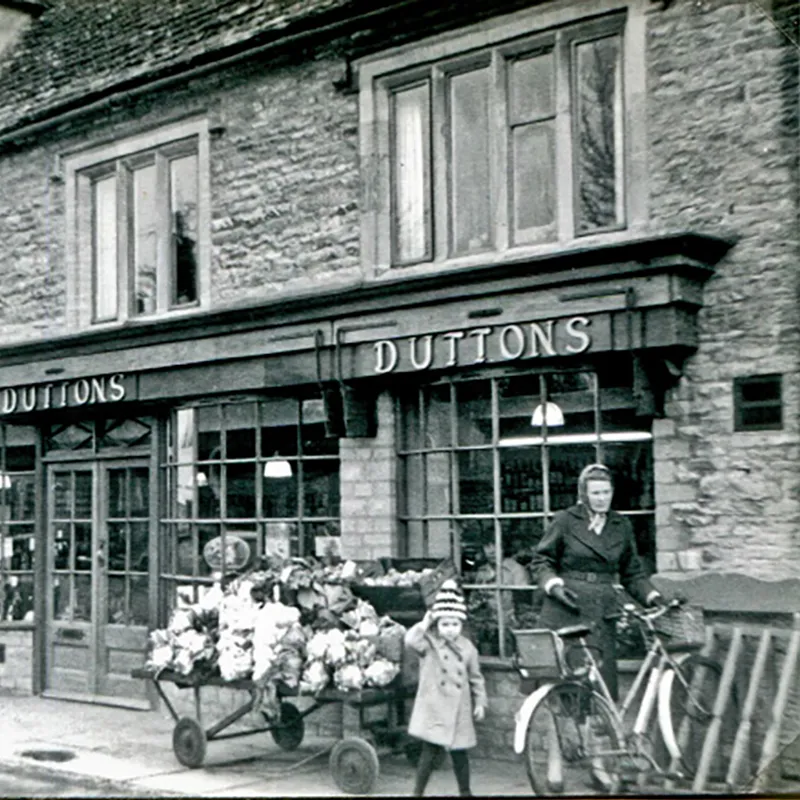 Black and white photograph of Duttons shop front with mother and daughter walking a bicycle.