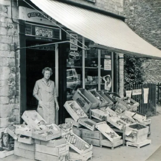 Vintage cool toned black and white photograph of lady outside a traditional greengrocers with crates of food outside.
