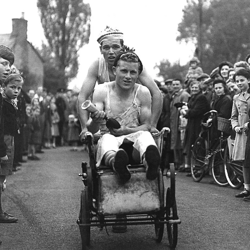 Vintage black and white photograph of two young men competing in a race, where one is pushing the other in a cart.
