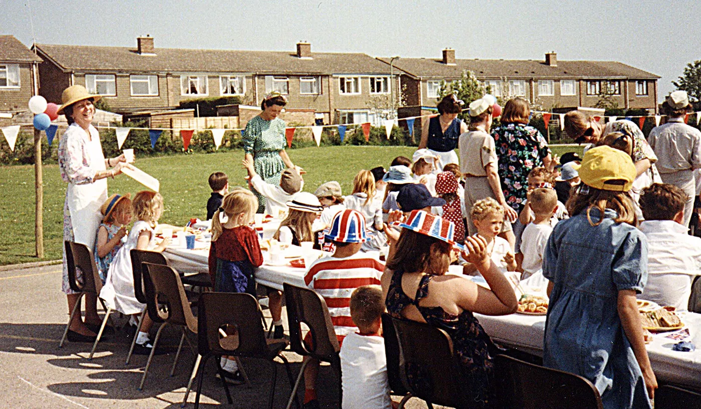 Black and white street party with people sat on benches and deck chairs along multiple tables pushed together